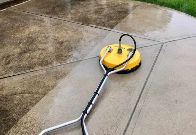 Yellow surface cleaner attached to a pressure washer cleaning a concrete patio, showing a clear contrast between cleaned and dirty sections.