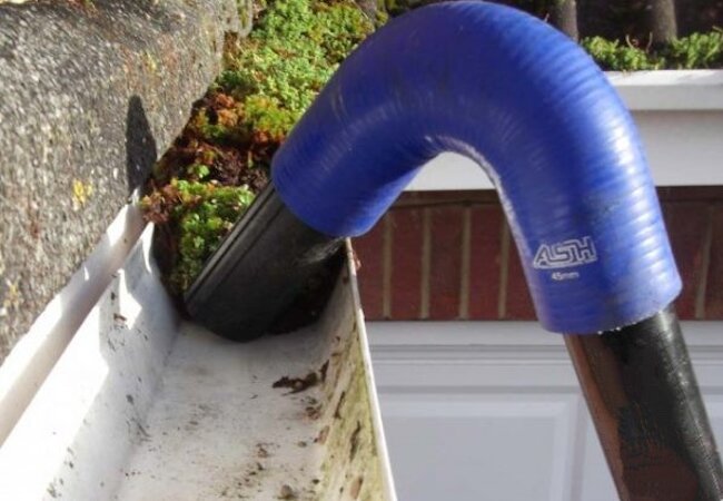 Blue flexible hose attached to a black tube cleaning debris and moss from a white gutter on a house.