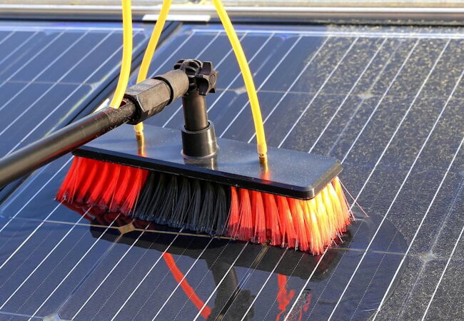 Cleaning brush with red and black bristles scrubbing dirt off a solar panel surface.