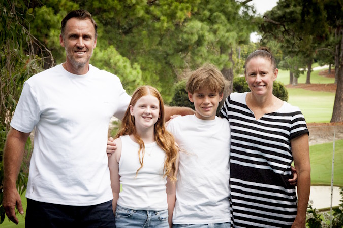 Family of four standing outdoors with trees and golf course in the background, smiling at the camera.